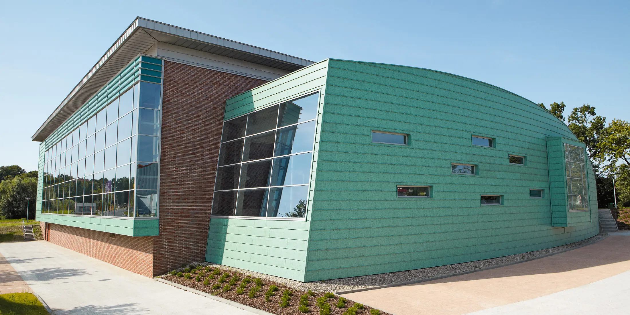 Modernes Schwimmbadgebäude mit auffälliger Fassade aus patinagrünen Aluminium-Paneelen und großen Glasflächen. Der Bau kombiniert gebogene und kantige Formen sowie unterschiedliche Materialien wie Metall und Backstein. Kleine, schmale Fenster lockern die Fassade auf. Das Gebäude steht in einer gepflegten Umgebung unter klarem, blauem Himmel.