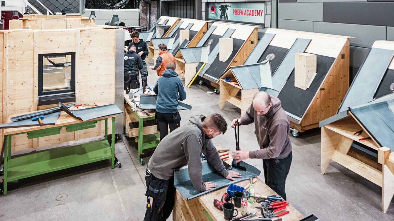 Einblick in die PREFA Academy, in der Verarbeiter an Dachmodellen arbeiten. Mehrere Auszubildende sind an Holztischen mit Dachmaterialien und Werkzeugen beschäftigt. Im Hintergrund sind die angefangenen Dachmodelle zu sehen.