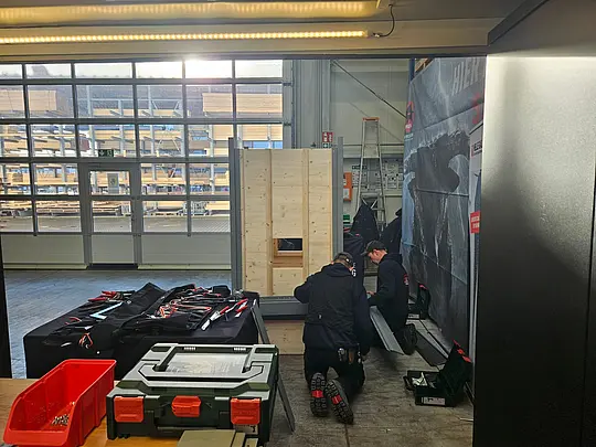 Praxisnahe Schulung mit PREFA Academy Cube: Zwei Handwerker montieren Sidings an ein Fassaden-Trainingsmodul in einer Werkhalle. Neben ihnen steht eine Arbeitsstation mit Spenglerwerkzeugen
