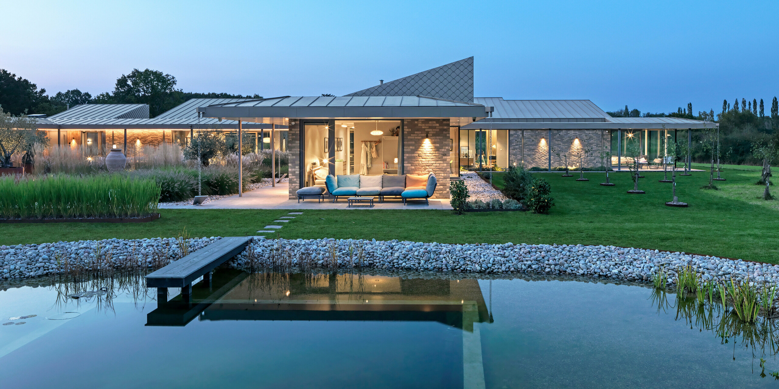 Abendansicht eines traumhaften Wohnhauses in West Sussex mit PREFALZ Aluminiumdach und Fassadenakzenten aus PREFA Wandrauten 20 × 20 in P.10 Bronze. Im Vordergrund ein angelegter Naturpool mit Holzsteg, dahinter eine großzügige Terrasse mit Loungemöbeln und beleuchteter Veranda. Die klare Linienführung der Architektur und die hochwertigen PREFA Produkte schaffen eine elegante Verbindung von Design, Funktionalität und Nachhaltigkeit.