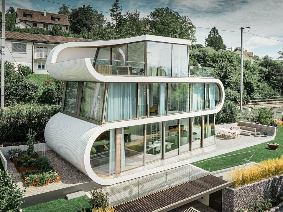 Aufnahme des modernen Flexhouses am Zürichsee. Mit seiner außergewöhnlichen Gebäudeform fällt das Flexhouse immer auf. Die Geschoßtrennungen ziehen sich als weißes Band geschwungen durch das Gebäude. Die runden Fassadenelemente wurden mit den Alu-Schindeln von PREFA in Reinweiß verkleidet.