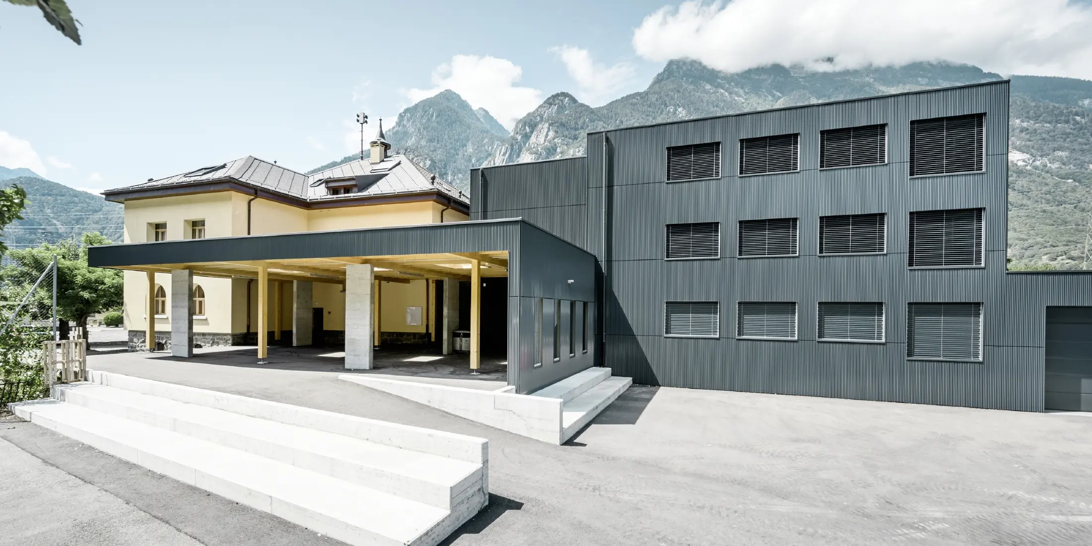 Schulgebäude mit einer Kombination aus historischem, gelb verputztem Altbau und modernem, anthrazitfarbenem Neubau mit Aluminiumfassade im Zackenprofil. Der Eingangsbereich ist überdacht und mit hellen Stützen aus Holz und Beton gestaltet. Im Hintergrund sind imposante Berge und ein teilweise bewölkter Himmel zu sehen.