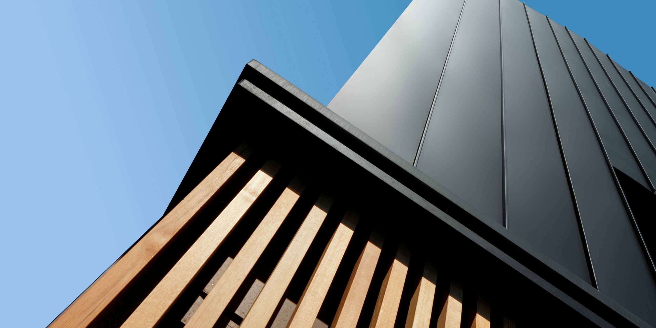 Architekturdetail eines modernen Doppelhauses in Cattolica: vertikale Aluminiumverkleidung mit PREFALZ in Schwarzgrau trifft auf elegante Holzlamellen. Die klare Linienführung und die hochwertige PREFA Oberfläche sorgen für Langlebigkeit, Korrosionsschutz und ein markantes Design unter strahlend blauem Himmel.