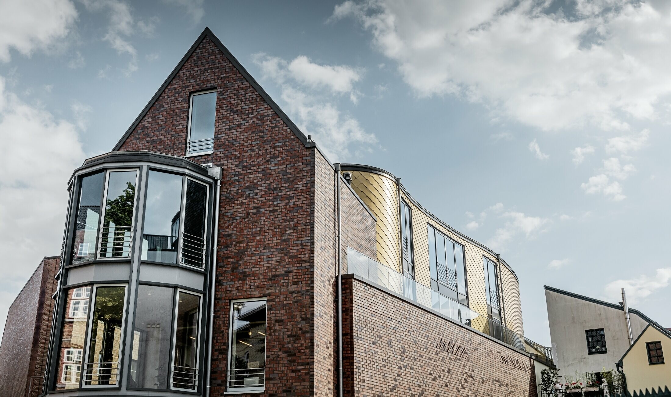 Geschäftshaus mit Backsteinfassade und großzügigen Glasflächen; Ein Teil der geschwungenen Fassade wurde mit der goldenen Wandraute aus Aluminium von PREFA verkleidet.