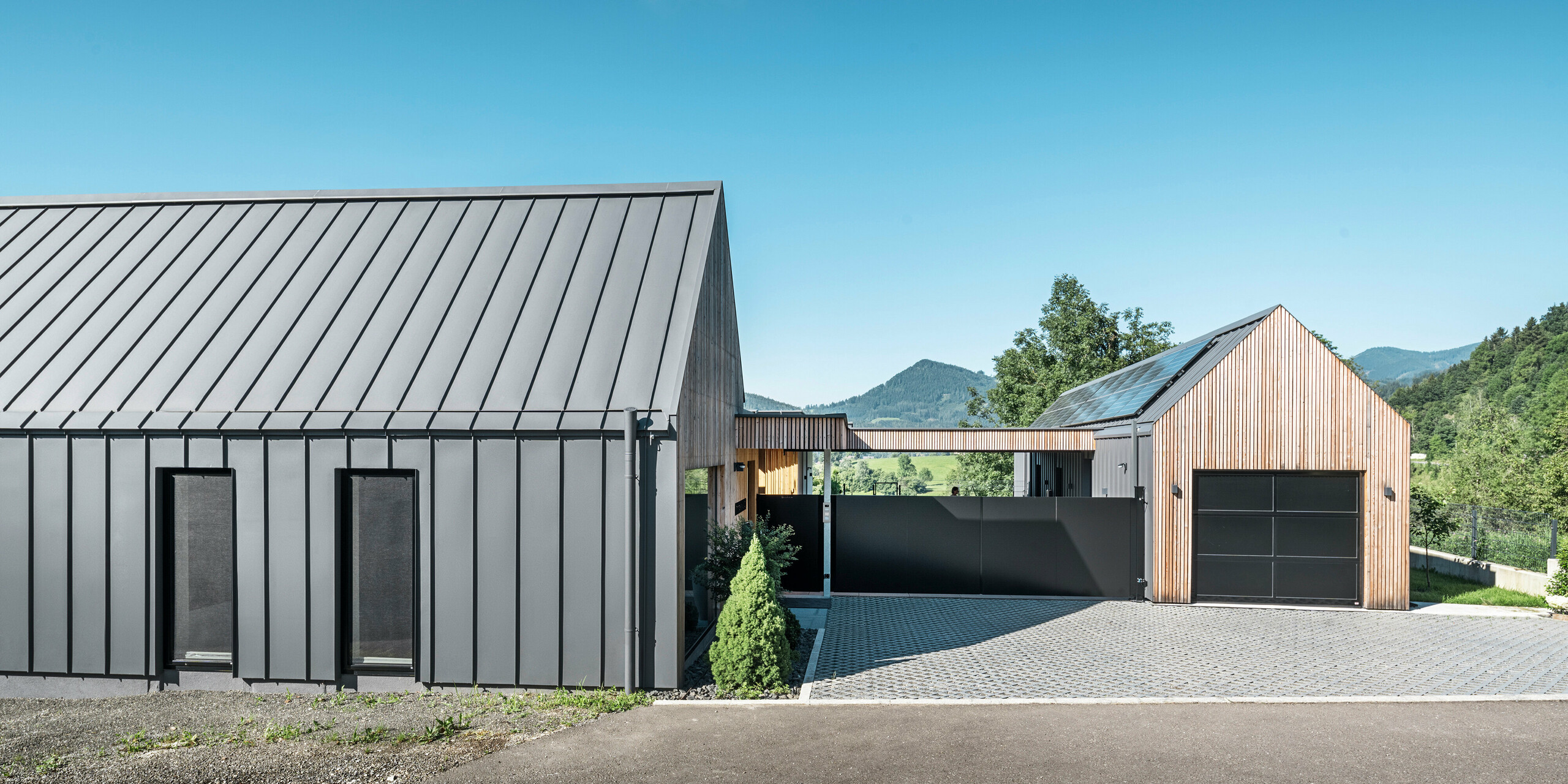 Vstupní areál rodinného domu s přilehlou garáží v rakouském Pogier uprostřed malebné krajiny. Střecha a fasáda obou budov jsou vybaveny odolným PREFALZ v P.10 tmavě šedé barvě, který vytváří moderní vzhled. Garáž se vyznačuje harmonicky integrovaným dřevěným obkladem, který hladce zapadá do okolní přírody. Dlážděné nádvoří a čisté linie designu doplňují estetiku nemovitosti, zatímco v pozadí zvlněné kopce dotvářejí obraz klidné zelené oázy v zeleném srdci Rakouska.