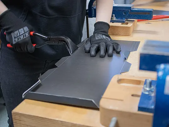 „Eine Schülerin arbeitet mit einer Zange an einem Aluminiumblech, das sie für das Vogelhaus bearbeitet. Das Blech ist bereits zugeschnitten und wird jetzt mit der Zange bearbeitet, um die gewünschte Form zu erreichen. Die Schülerin trägt Schutzhandschuhe und konzentriert sich auf die Fertigung des Teils. Im Hintergrund sind Werkzeuge und eine Werkbank zu sehen.