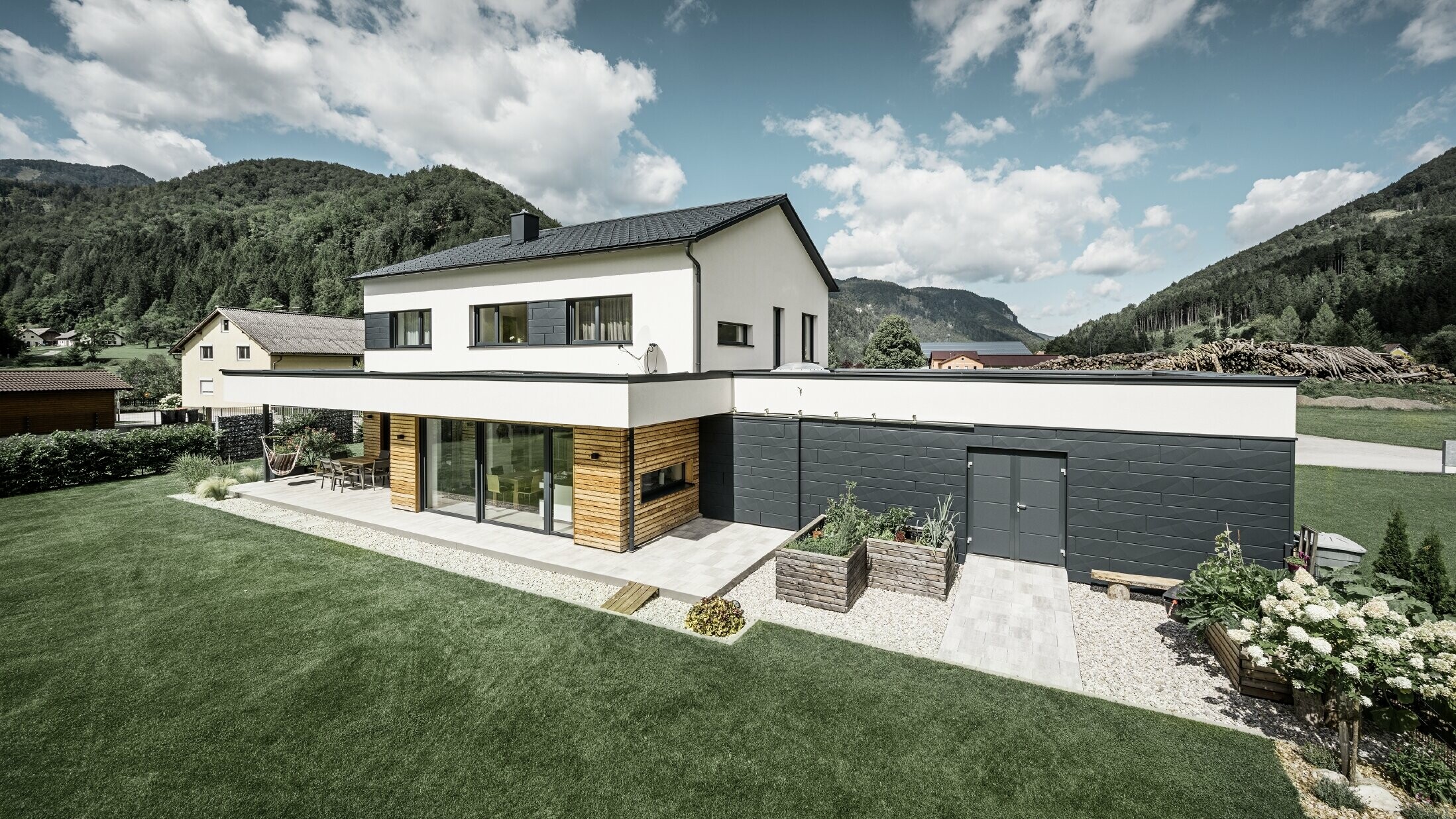 Das Bild zeigt ein modernes Einfamilienhaus in einer idyllischen Landschaft mit einer klaren Sicht auf bewaldete Hügel im Hintergrund. Das Haus zeichnet sich durch eine Kombination aus weißen Putzwänden und einer markanten Fassadenverkleidung mit PREFA Siding.X in P.10 Anthrazit aus, die für einen modernen und langlebigen Look sorgt. Die dunkle Färbung der Fassadenelemente bildet einen ansprechenden Kontrast zum hellen Außendesign und zur natürlichen Umgebung. Die Anordnung der Siding.X Fassadenpaneele ist präzise und verleiht der Immobilie einen strukturierten und hochwertigen Charakter, der typisch für PREFA Produkte ist. Das Dach des Gebäudes wurde mit der bewährten PREFA Dachplatte eingedeckt. Die Dachentwässerung erfolgt durch das Ablaufrohr und die Hängerinne von PREFA.