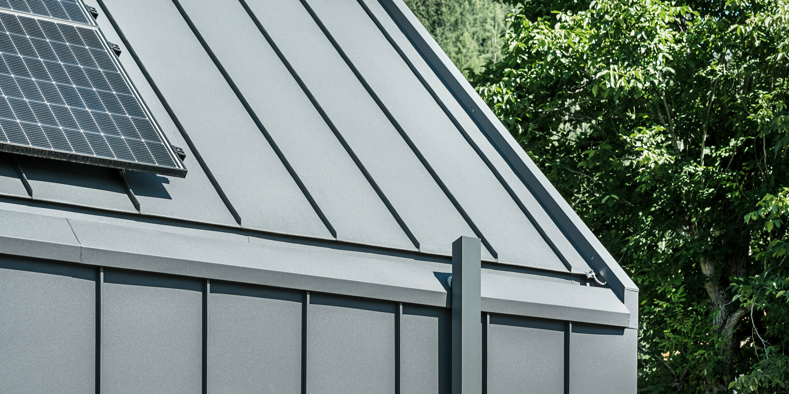 Detailní záběr střechy nebo fasády moderního rodinného domu v rakouském Pogier s udržitelným a trvanlivým pláštěm budovy od PREFA. Střecha a fasáda jsou opláštěny PREFALZ v barvě P.10 tmavě šedá, což zajišťuje elegantní a nadčasový vzhled. Dům je navíc vybaven kvalitním odvodněním střech, které v architektuře dokonale spojuje funkčnost a estetiku. Dešťová voda je odváděna čtvercovými trubkami PREFA, které jsou – podobně jako střecha a fasáda – rovněž v tmavě šedé barvě.Na levém okraji obrázku je na střeše vidět fotovoltaický systém, který dále demonstruje udržitelný koncept budovy. V pozadí jsou vidět zelené stromy a modrá obloha.