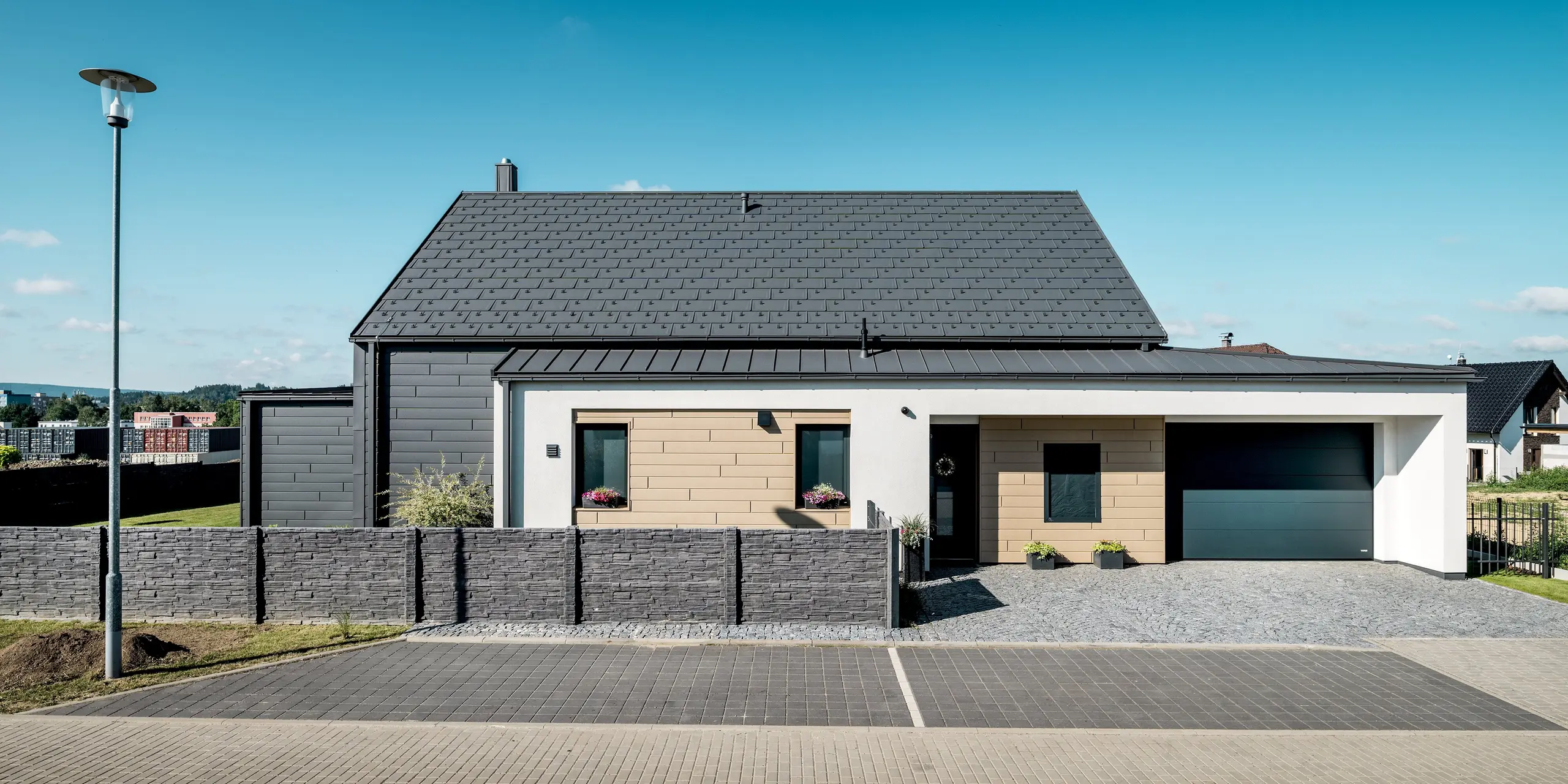Frontansicht des modernen Einfamilienhauses mit PREFA Aluminiumlösungen an Dach und Fassade. Die PREFA Dachplatte R.16 in P.10 Dunkelgrau sowie die Sidings in P.10 Dunkelgrau und P.10 Sandbraun schaffen ein harmonisches Zusammenspiel aus klaren Linien und zeitgemäßem Design. Die langlebige Aluminiumhülle schützt das Gebäude zuverlässig und verleiht ihm eine elegante, dauerhaft ansprechende Optik.