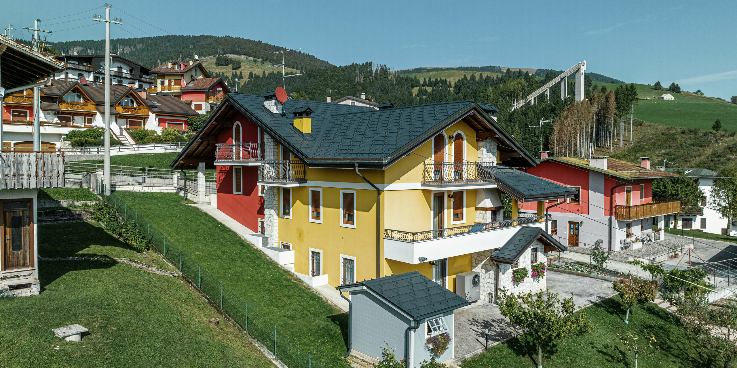 Einfamilienhaus in Gallio in traditioneller Alpenarchitektur mit leuchtend gelb-roter Putzfassade und Natursteinelementen. Das Dach aus PREFA Dachplatten R.16 in P.10 Dunkelgrau bietet höchste Witterungsbeständigkeit und harmoniert perfekt mit der Umgebung. Die Dachentwässerung mit PREFA Hängerinne und Ablaufrohr - ebenfalls in P.10 Dunkelgrau - sorgt für zuverlässigen Schutz. Umgeben von grünen Wiesen und mit Blick auf die Berge vereint das Haus Ästhetik, Langlebigkeit und Funktionalität. Im Hintergrund ist die bekannte Skisprungschanze der Region zu erkennen.