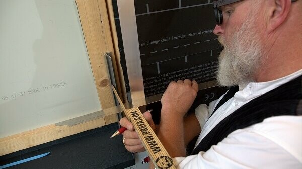 Techniker Tom Weiß nimmt mit einem Maßstab das Schenkelmaß eines F-Profils (Flutzprofils), dass er seitlich an einer Holz-Fensterlaibung angebracht hat. 
