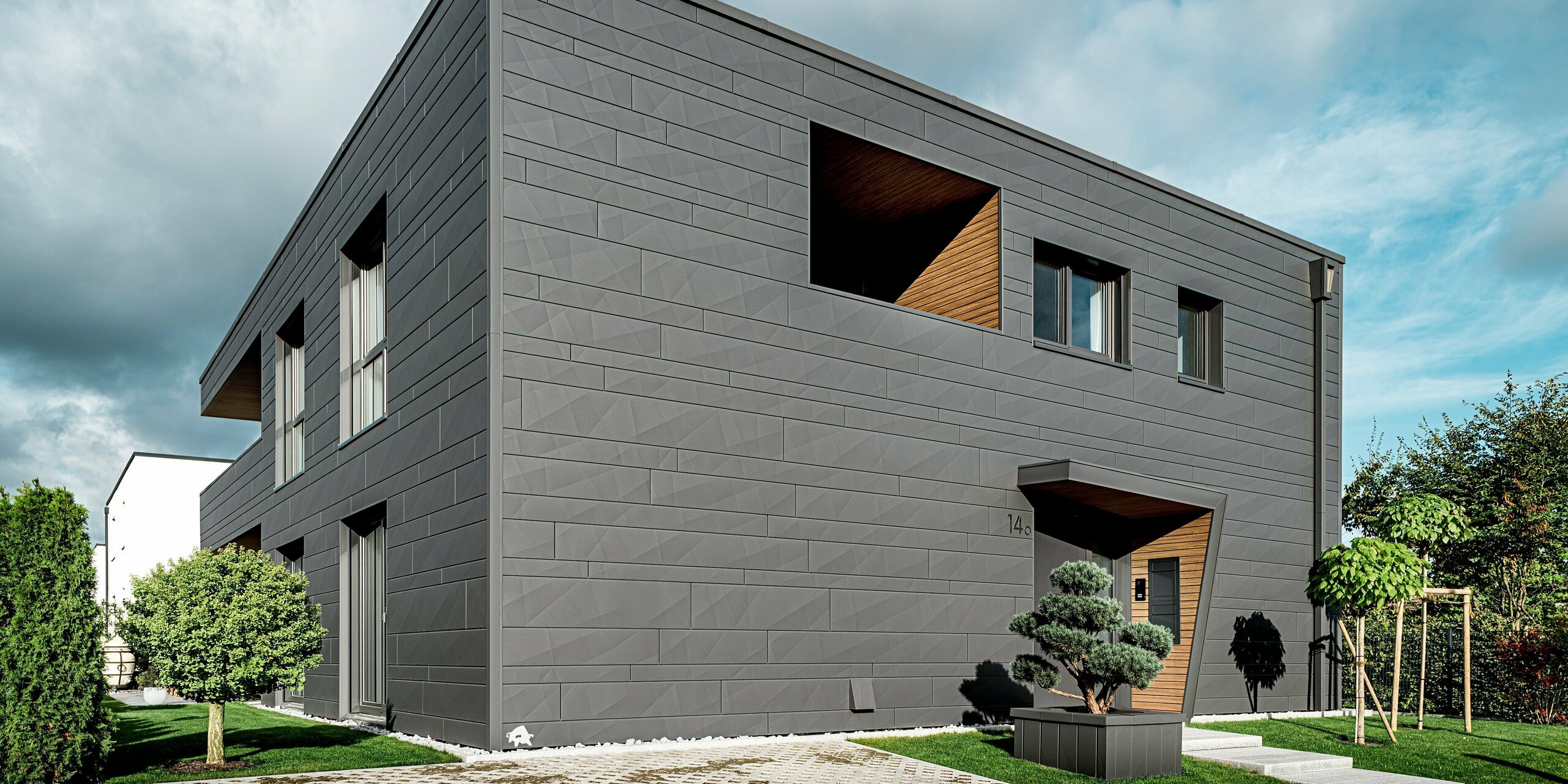 Das moderne Einfamilienhaus in Heidenheim zeigt eine vollständig mit PREFA Siding.X in P.10 Dunkelgrau verkleidete Aluminiumfassade, deren markante Kantungen und unterschiedlichen Paneelbreiten dem klaren Baukörper eine dynamische Struktur verleihen. Der holzverkleidete Eingangsbereich bildet einen warmen Kontrast zur eleganten Metalloberfläche. Im Vordergrund steht eine sorgfältig gestaltete Gartenfläche. Die robuste PREFA Aluminiumfassade und das integrierte Quadratrohr bieten langlebigen Witterungsschutz, hohe Stabilität und ein zeitlos modernes Erscheinungsbild.