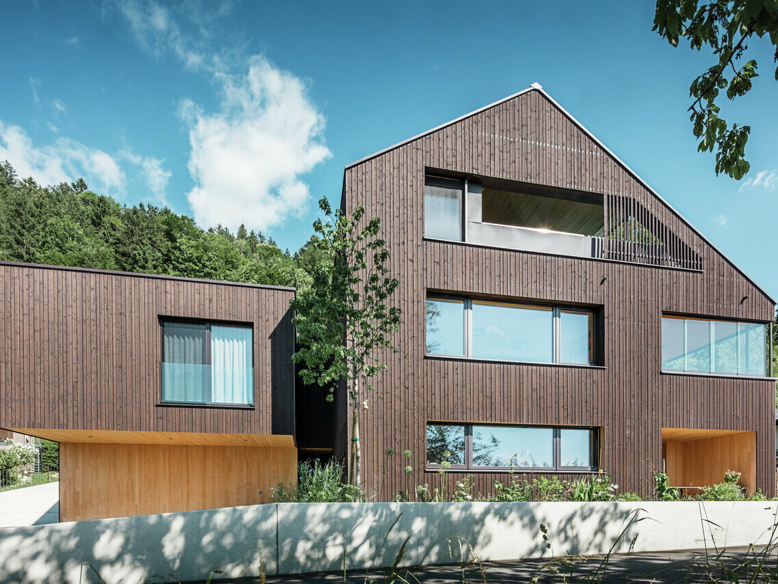 Ein modernes Mehrfamilienhaus mit nussbraunem PREFA Prefalz Dach und dunkler Holzfassade, durchsetzt mit hellen Holzelementen, großen Fenstern und umgeben von grüner Landschaft.
