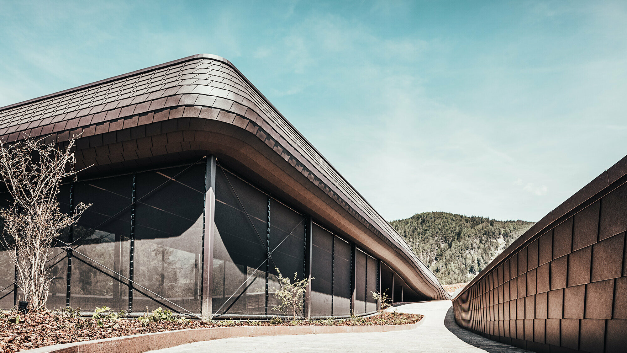 Seitliche Ansicht des Außenbereiches des Hotels in Südtirol. Die PREFA Wandschindeln sind an jeder Fläche zu sehen. Im Hintergrund ist die hügelige, bewaldete Landschaft zu sehen.