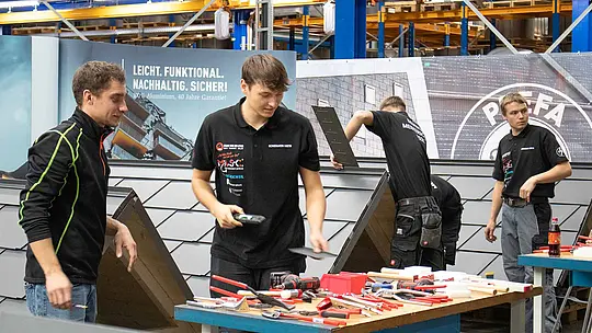 Die teilnehmenden Meisterschüler der Deutschen Meisterschaft im Klempnerhandwerk 2025 arbeiten in der PREFA Academy in Wasungen an Übungsmodellen mit Aluminiumdächern. Auf den Arbeitstischen liegen Werkzeuge und Materialien bereit. Im Hintergrund sind PREFA-Banner und Werkstatteinrichtungen zu sehen.