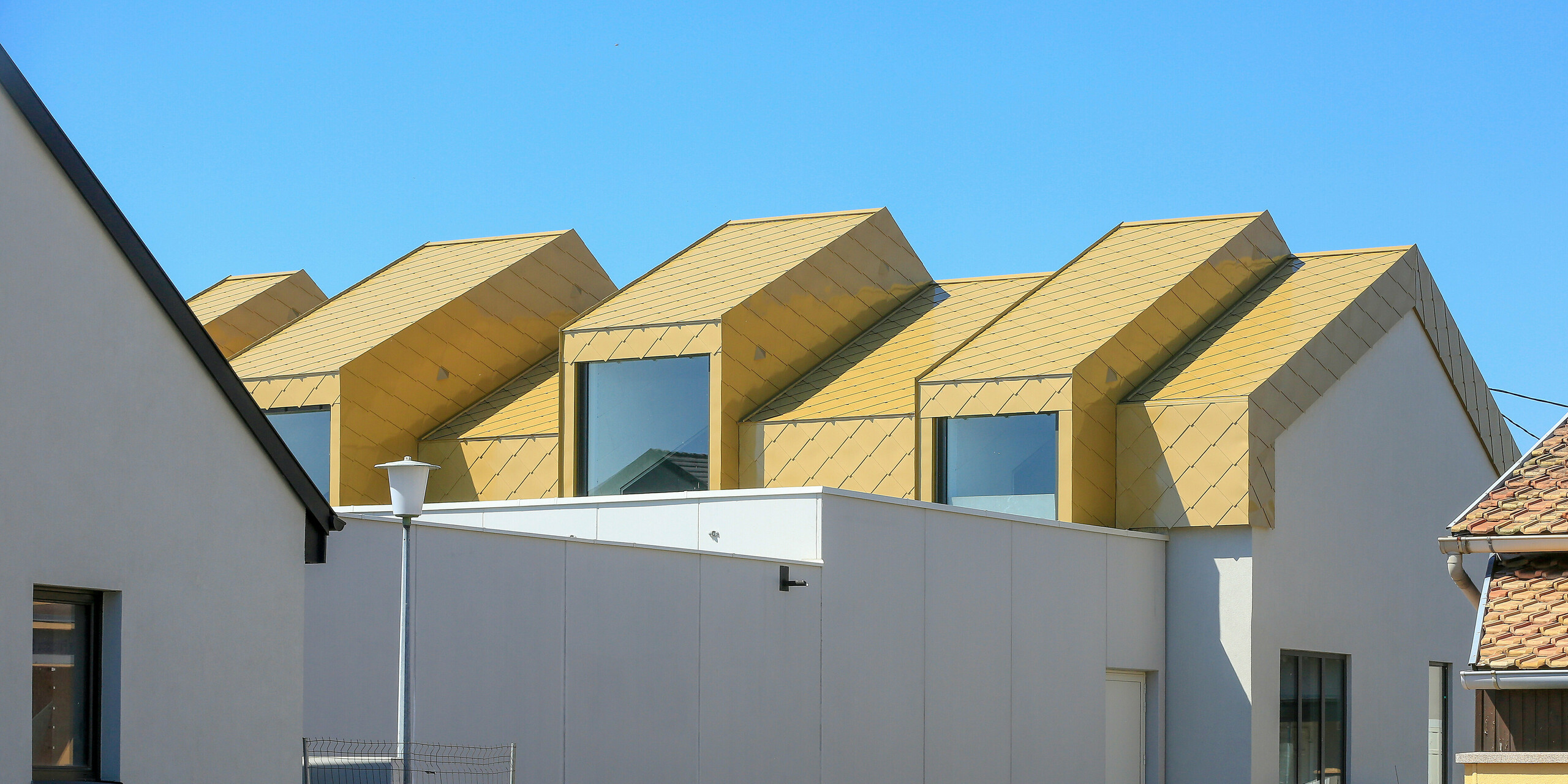 Blick aus der Distanz auf das Gemeinde- und Kulturzentrum Fessenheim (FRA). Die Perspektive auf das Gebäude mit den glänzenden PREFA Dach- und Wandrauten 44 × 44 in Sparkling Gold ist von der Straße.