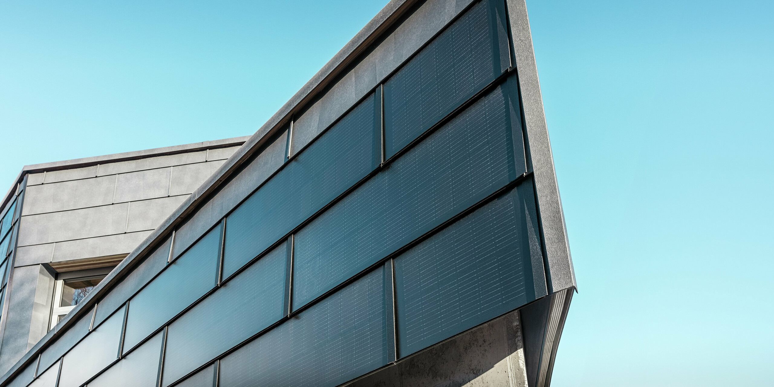 Moderne Firmenfassade der Spenglerei RÜMA in Vorarlberg mit PREFA Solardachplatten aus Aluminium und integrierten Photovoltaikmodulen, kombiniert mit PREFA Fassadenpaneelen FX.12 für nachhaltige Energiegewinnung und dynamisches Fassadendesign. Die Aufnahme zeigt die Gebäudeecke aus nächster Nähe. Im Hintergrund ist der strahlend blaue Himmel zu sehen. 