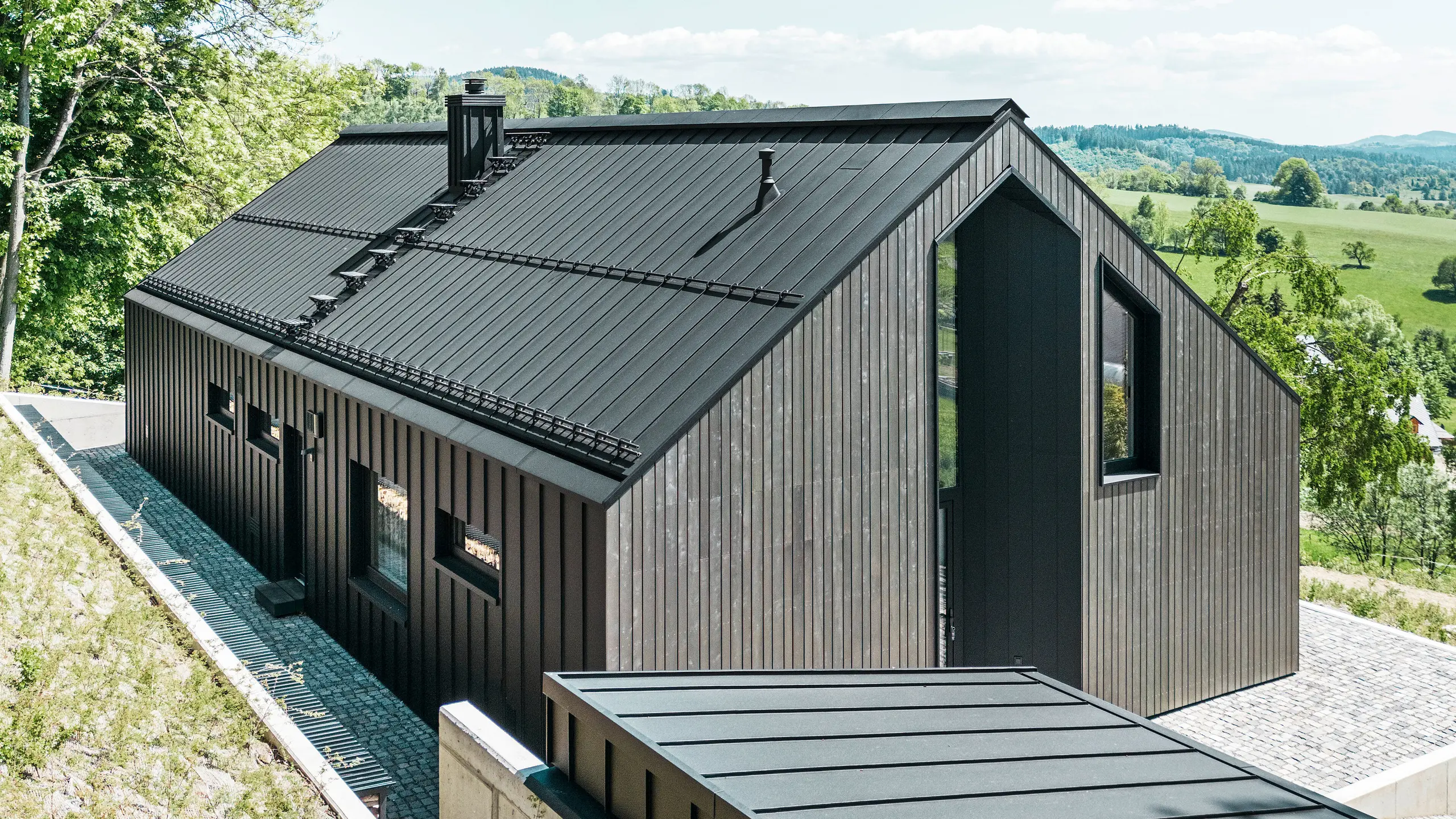 A dark solid-timber house clad in PREFALZ in P. 10 on the roof and the reverse of the building. In front of the house stands a small outbuilding with an aluminium cladding of PREFALZ in P.10 black. In the background, a bright blue sky and woodland.