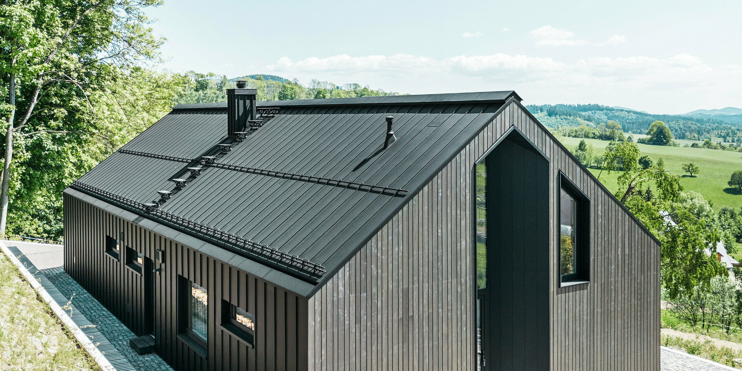 Dunkles Holzmassivhaus mit PREFALZ in P. 10 am Dach und der Rückseite des Gebäudes. Vor dem Haus steht ein kleines Nebengebäude mit einer Aluminiumhülle aus PREFALZ in P.10 Schwarz. Im Hintergrund ein strahlend blauer Himmel und Wald.