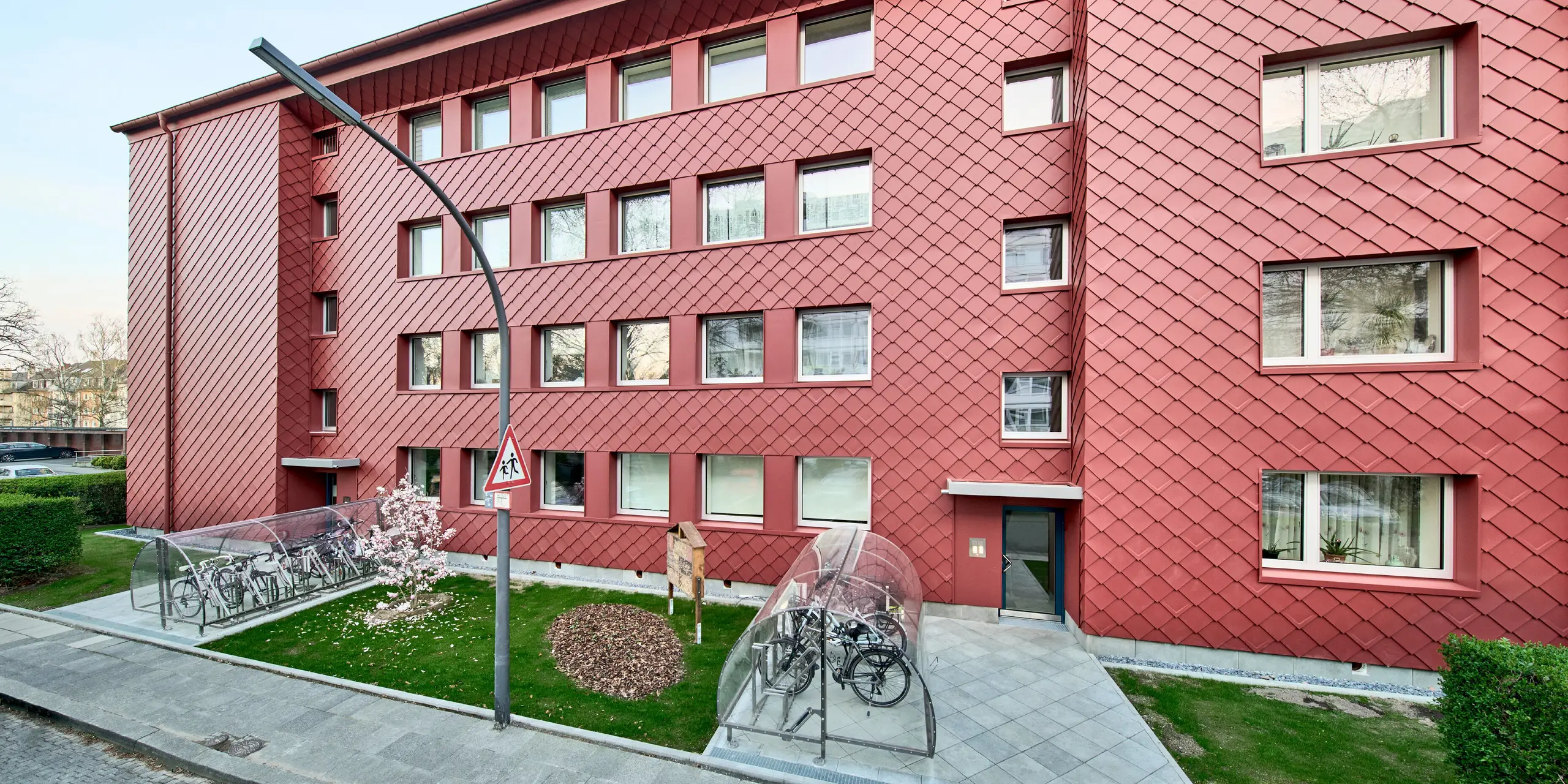 Markante Architektur in P.10 Oxydrot: Das sanierte Mehrfamilienhaus „Energiesprong 2426“ in Köln-Zollstock begeistert mit einer ausdrucksstarken Fassade aus PREFA Wandrauten 29 × 29. Das dynamische Rautenmuster, geometrisch angeordnete Fenster und farblich abgestimmte Dachentwässerung prägen das moderne Erscheinungsbild. Die leichte, langlebige und witterungsbeständige Aluminiumverkleidung vereint nachhaltige Sanierung mit anspruchsvollem Design.
