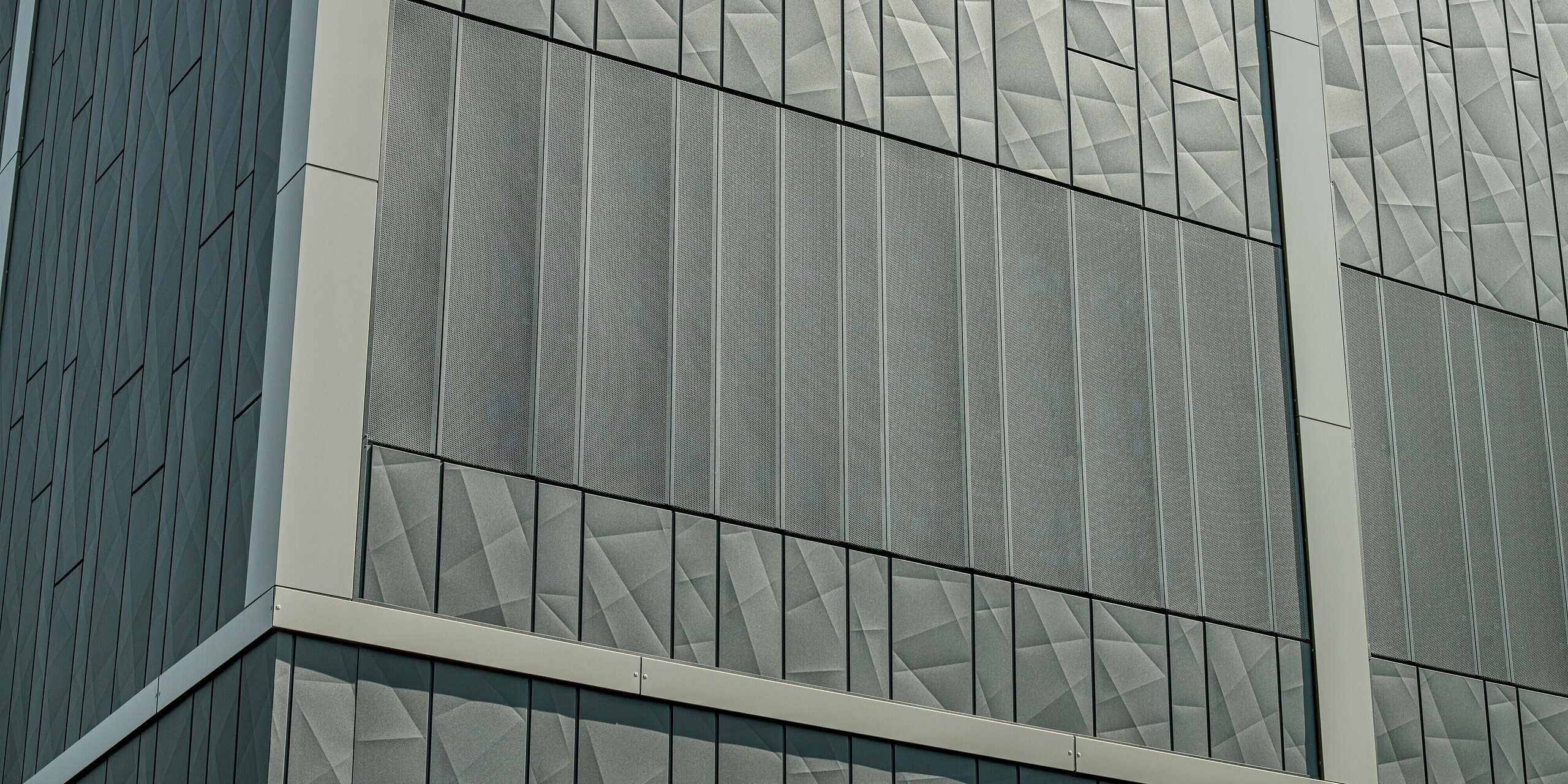 Das Bild zeigt einen detailreichen Ausschnitt der Aluminiumfassade des Einkaufszentrums in Rohrbach. Besonders auffällig sind die anthrazitfarbenen Siding.X-Paneele mit ihrer geometrischen Struktur und den variierenden Oberflächen, die einen dynamischen, modernen Eindruck vermitteln.  Im unteren Bereich des Bildes sind perforierte Siding-Elemente zu sehen. Diese setzen nicht nur ästhetische Akzente, sondern sorgen auch für eine gezielte Belüftung und Lichtdurchlässigkeit. Die Kombination aus glatten und perforierten Paneelen verleiht der Fassade somit eine lebendige und funktionale Gestaltung, die optisch und praktisch überzeugt.