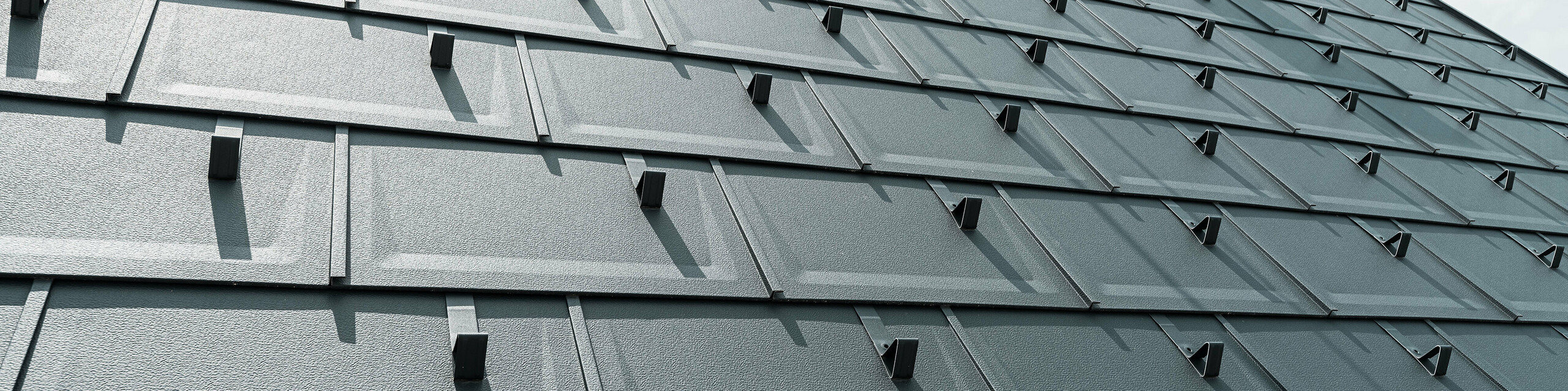 Detailaufnahme der PREFA Dachplatte R.16 in der Farbe P.10 Anthrazit. Am Dach sind PREFA Schneestopper und ein Rauchfang zu sehen.