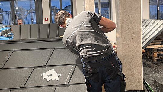 Ein Teilnehmer der Deutschen Meisterschaft im Klempnerhandwerk richtet das Metallblech exakt aus. Der Wettbewerbsbereich ist von modernen Arbeitsplätzen und Maschinen umgeben, und der Fokus liegt auf präzisem Handwerk.