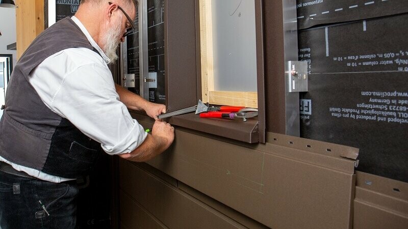 Zu sehen ist ein PREFA Experte, der an der Fassade unter einem Fenster arbeitet. Konkret klinkt er hier die PREFA Siding Fassadenplatten mit geringem Aufwand aus.