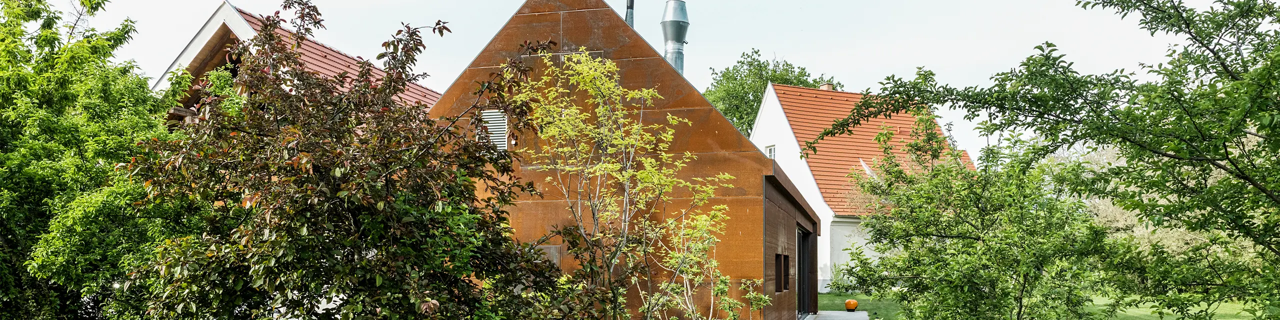 Wohnhaus Velich im Seewinkel, Burgenland: Blick auf die moderne Gartenanlage mit schönen Pflanzen. Das Wohnhaus dient als Veranstaltungsort für den Fachdialog PREFARENZEN 2025, bei dem Architekten und PREFA-Experten die interessantesten Architekturobjekte auswählen. Im Vordergrund ist eine stilvolle, rostfarbene Metallwand mit der Aufschrift „RESIDENCE VELICH“ zu sehen. Die Architektur des Gebäudes verbindet traditionelle und moderne Elemente und fügt sich harmonisch in die natürliche Umgebung ein.