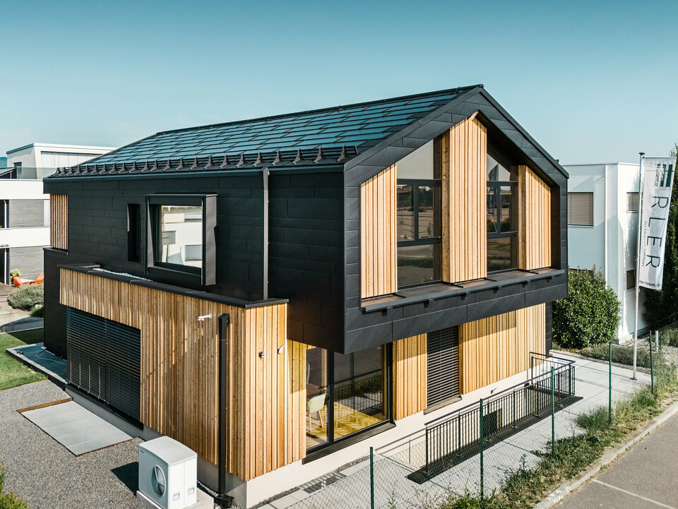 Vorderseite des Musterhauses Autark im Fertighauszentrum Blaue Lagune, Vösendorf (AT). Das Dach ist mit schwarzen PREFA Solardachplatten und Dachplatten R.16 in hochwertiger P.10-Beschichtung eingedeckt. Die Fassade besteht aus einer Kombination von vertikalen Holzelementen und PREFA Siding.X in P.10 Schwarz, was dem Gebäude einen modernen und nachhaltigen Charakter verleiht. Am rechten Bildrand weht die Fahne der Erler Bau GmbH.