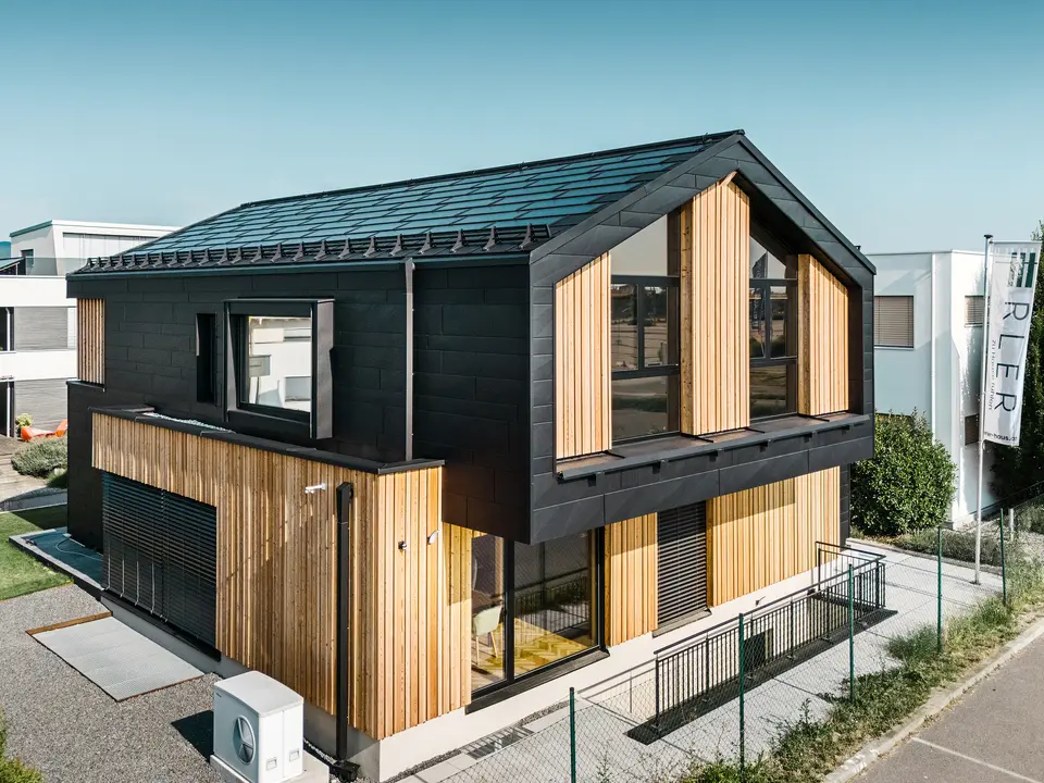 Vorderseite des Musterhauses Autark im Fertighauszentrum Blaue Lagune, Vösendorf (AT). Das Dach ist mit schwarzen PREFA Solardachplatten und Dachplatten R.16 in hochwertiger P.10-Beschichtung eingedeckt. Die Fassade besteht aus einer Kombination von vertikalen Holzelementen und PREFA Siding.X in P.10 Schwarz, was dem Gebäude einen modernen und nachhaltigen Charakter verleiht. Am rechten Bildrand weht die Fahne der Erler Bau GmbH.