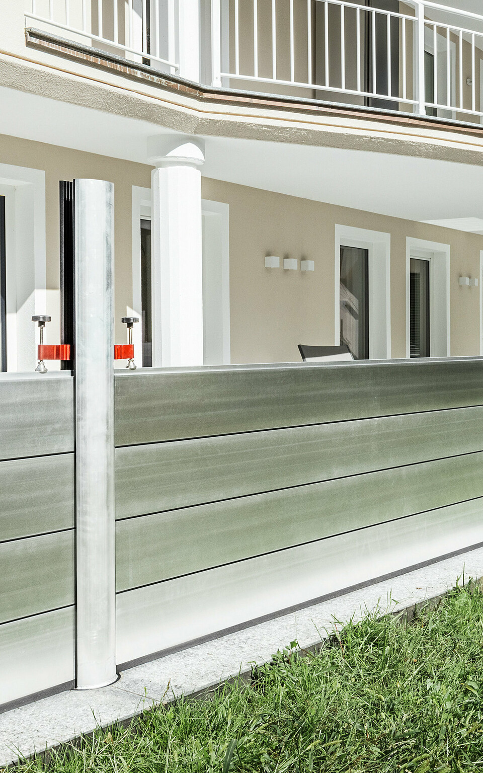 Panoramaansicht eines exklusiven Wohnhauses in Tirol mit installiertem PREFA Hochwasserschutzsystem entlang der Terrasse. Die Dammbalken aus Aluminium verlaufen über die gesamte Gebäudelänge und schützen mehrere Zugänge wie Terrassentüren und Fenster vor möglichem Hochwasser aus dem angrenzenden Gelände. Die moderne Architektur des Hauses mit Rundbalkon und weißen Stützen bildet einen stilvollen Kontrast zum funktionalen Hochwasserschutz aus Aluminium.