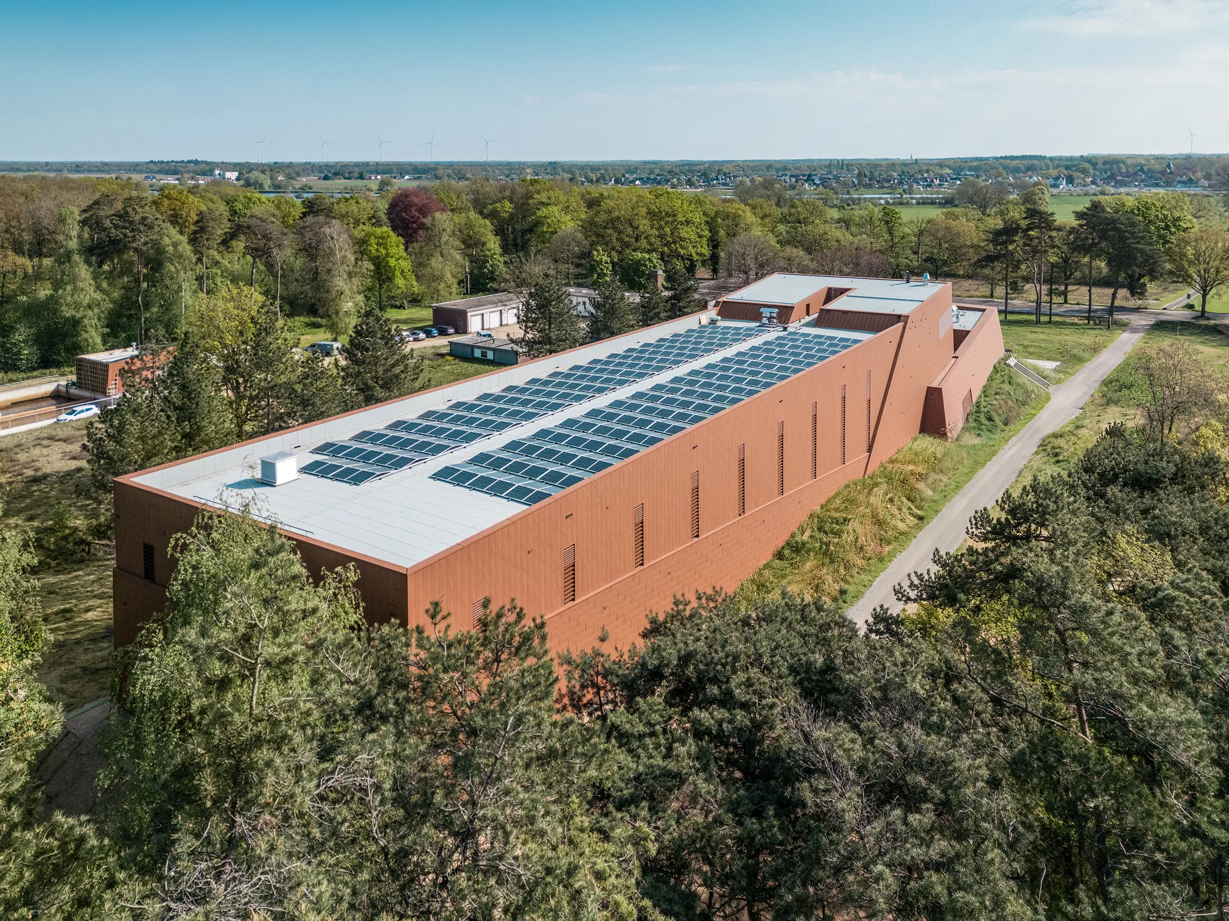 Luftaufnahme des Trinkwasserpumpwerks WML in Lottum, eingebettet in eine weitläufige Landschaft aus Bäumen und Feldern. Die langgestreckte Gebäudestruktur beeindruckt mit einer prägnanten PREFA Siding.X Aluminiumfassade in warmer Rostoptik und einem Dach voller Photovoltaikmodule als nachhaltigem Eyecatcher. Die fein strukturierte Gebäudehülle reflektiert Licht lebendig und unterstreicht die moderne Formensprache. Die leichten, langlebigen und recycelbaren PREFA алюминиumpaneele garantieren hohe Witterungsbeständigkeit, Energieeffizienz und eine zukunftsorientierte, wartungsarme Architektur.