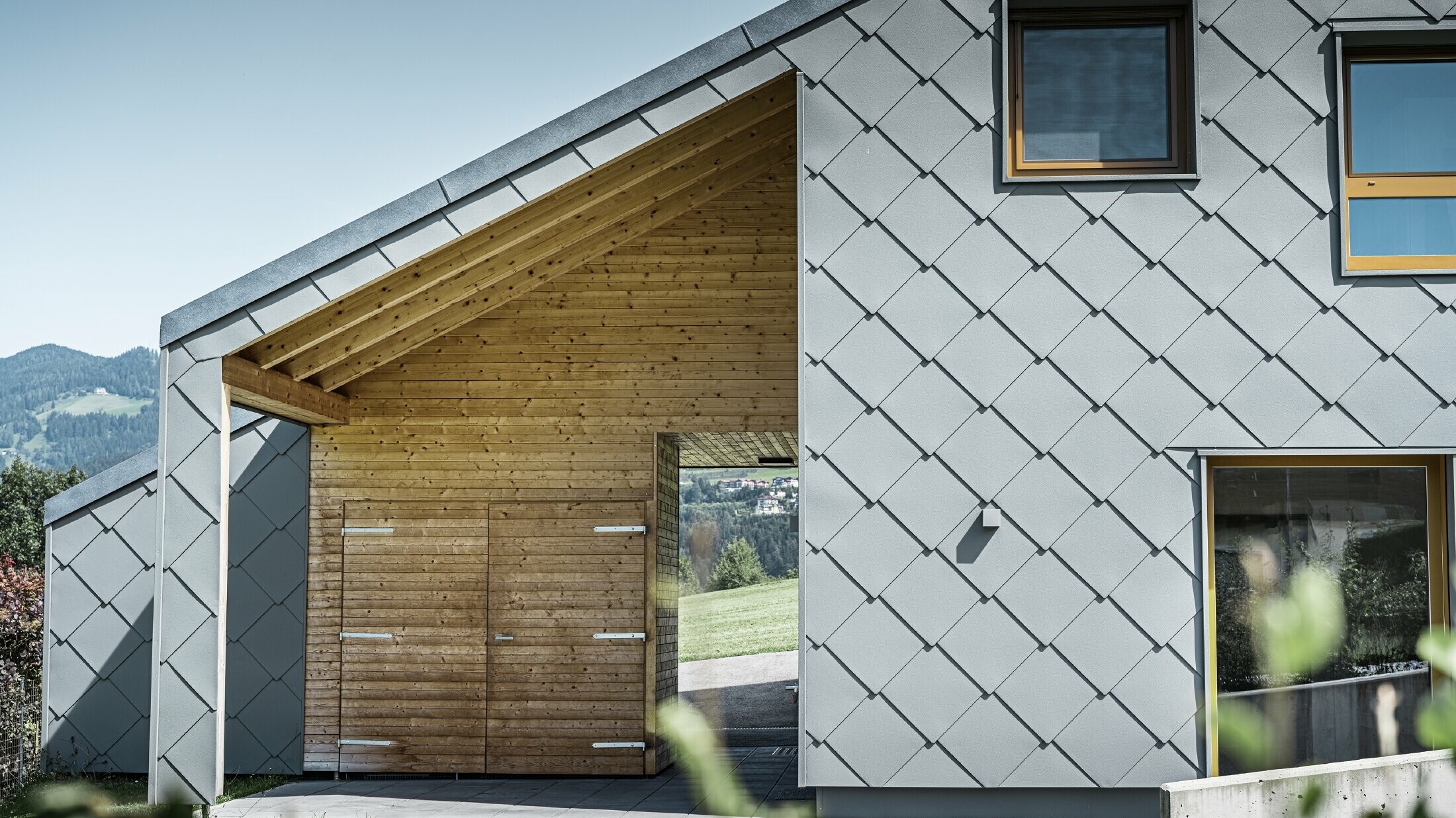 Der überdachte Eingangsbereich ist mit waagrechter Holzverkleidung gemacht, der Rest der Fassade ist mit der großen PREFA Wandraute aus Aluminium in Hellgrau verkleidet