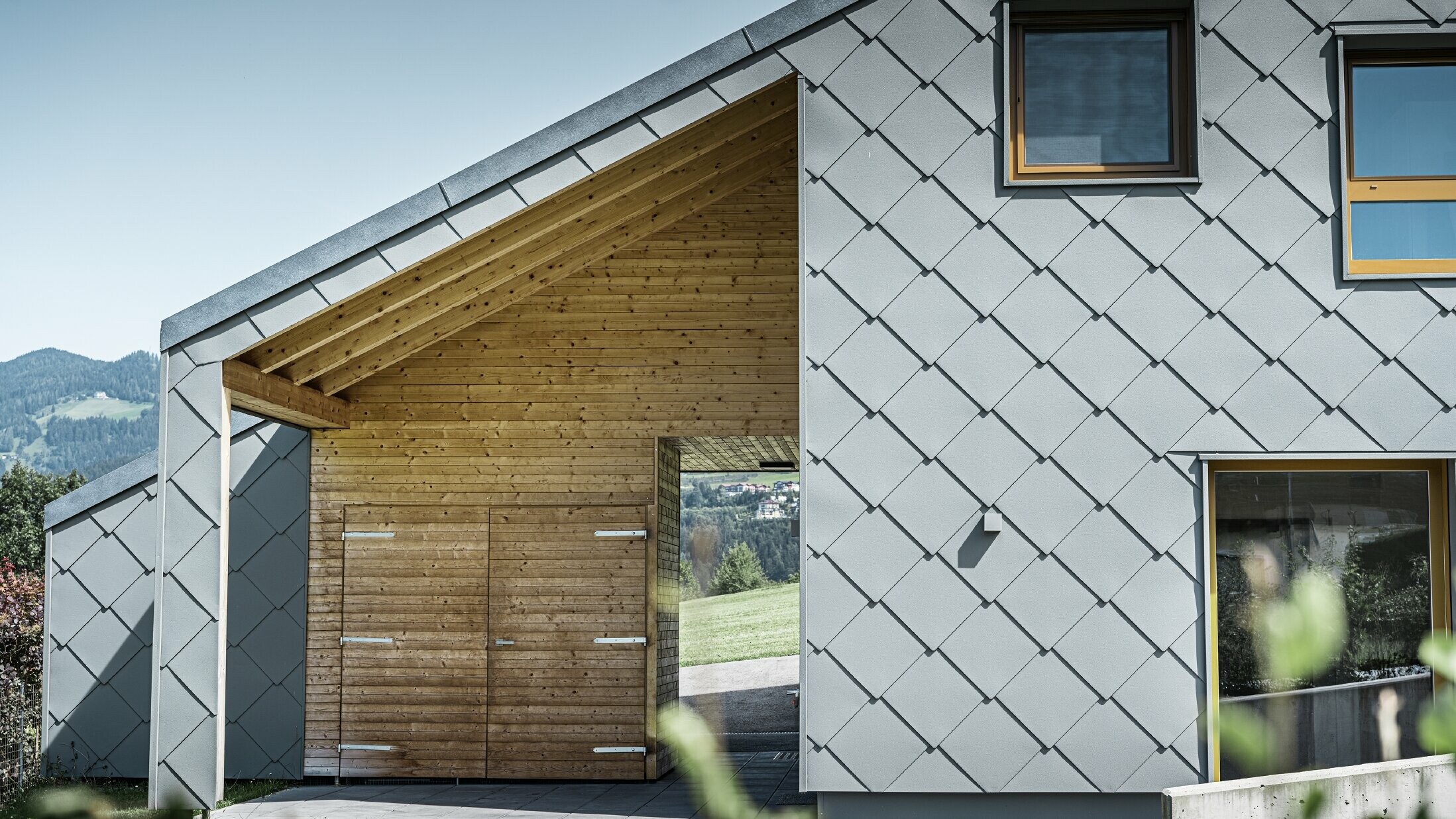 Der überdachte Eingangsbereich ist mit waagrechter Holzverkleidung gemacht, der Rest der Fassade ist mit der großen PREFA Wandraute aus Aluminium in Hellgrau verkleidet