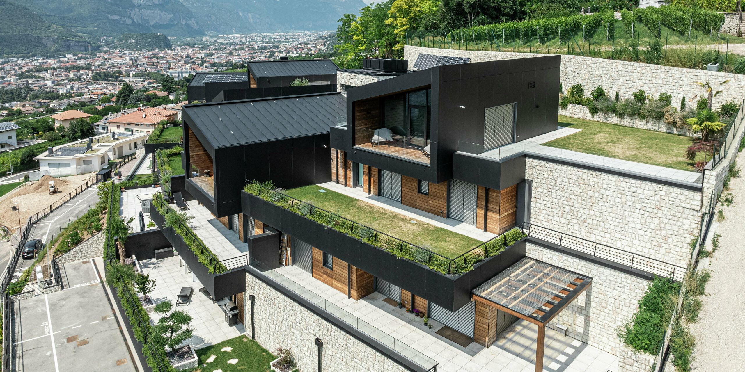 Exklusive Wohnanlage mit Panoramablick über Trient: Die moderne Architektur fügt sich harmonisch in die Hanglage ein und kombiniert hochwertige Materialien wie Naturstein, Holz und Aluminium. Die Fassade aus PREFA Aluminium Verbundplatten in P.10 Schwarz sorgt für eine edle und langlebige Optik, während das PREFALZ Dach in P.10 Schwarz optimalen Schutz vor Witterungseinflüssen bietet. Grunflächen und großzügige Terrassen unterstreichen das nachhaltige Wohnkonzept und schaffen eine perfekte Verbindung von Natur und modernem Design.