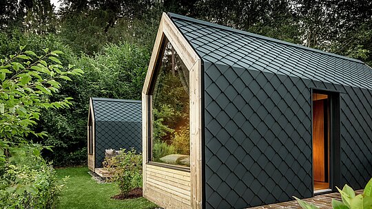 Zwei minimalistische Tiny Houses des Secret Koi Garden stehen eingebettet im satten Grün am Natterer See. Die Fassaden und Dächer sind vollständig mit kleinteiligen PREFA Aluminiumrauten in P.10 Anthrazit verkleidet und erzeugen eine markante, fischhautartige Struktur. Große Glasflächen und warme Holzdetails bilden einen spannenden Kontrast zum dunklen Aluminium. Das langlebige, witterungsbeständige PREFA Aluminium schützt zuverlässig und unterstreicht die hochwertige Glamping‑Architektur.