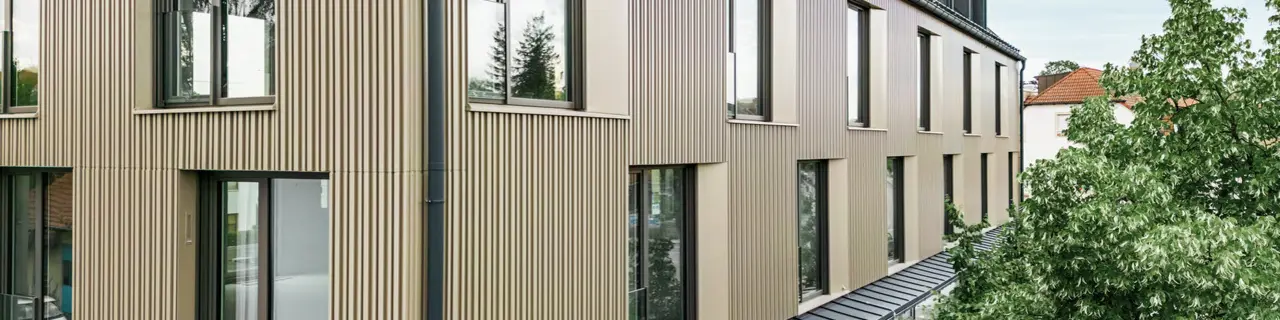 Das Bild zeigt ein modernes Gebäude in Feldkirch, entworfen vom Architekten Stefan Heigl. Die Fassade ist aus Prefalz in der Farbe Bronze gestaltet, und das Satteldach ist mit Prefalz in Schwarzgrau gedeckt. Große Fenster sorgen für viel Licht im Inneren, während die verglaste Erdgeschosszone eine einladende Gastronomiefläche bietet. Die hochwertige Materialwahl und das elegante Design verleihen dem Gebäude ein zeitgemäßes und stilvolles Erscheinungsbild.