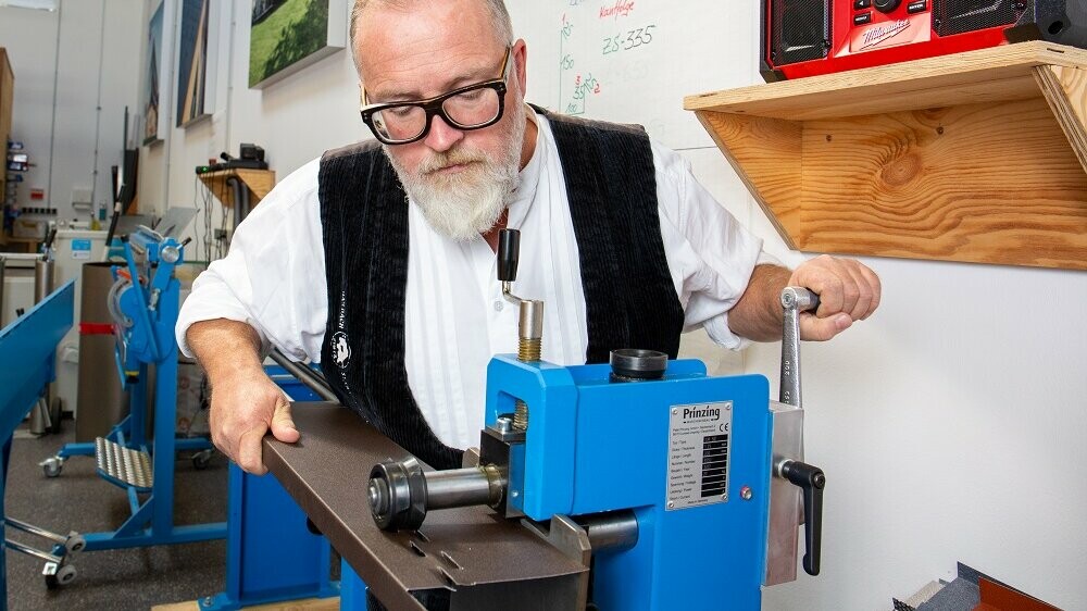 Auf diesem Bild ist neben dem PREFA Techniker eine blaue Maschine zu erkennen. Mithilfe dieser Maschine stanzt er die Zuluft-Löcher in das Sturzblech.