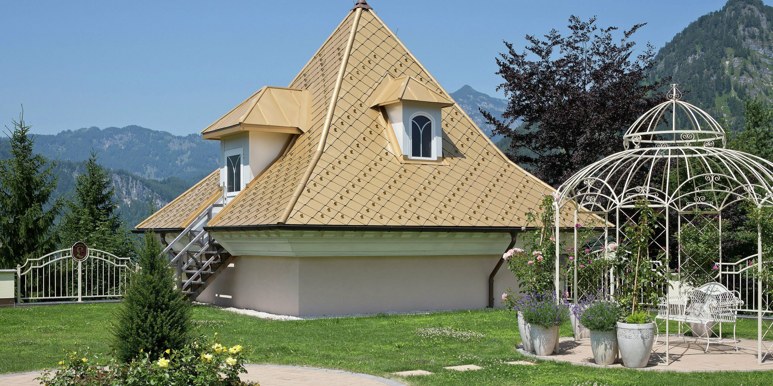 Kleiner Zubau eines Einfamilienhauses in Tirol mit PREFA Dachrauten in Mayagold und einem malerischen Garten im Vordergrund, umgeben von den Alpen.