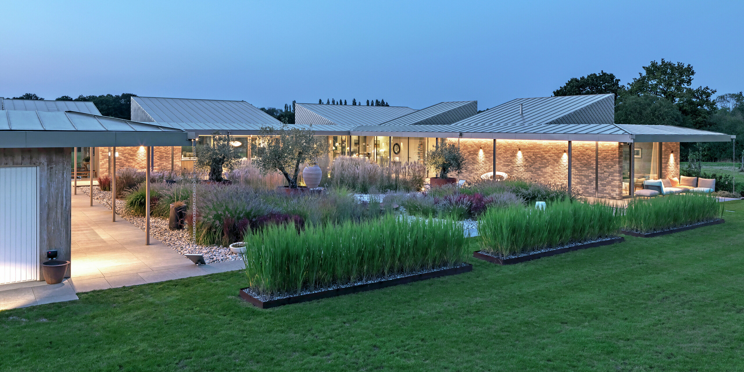Modernes Wohnhaus in West Sussex mit PREFALZ Aluminium Stehfalzdeckung und PREFA Wandrauten 20 × 20 aus P.10 Bronze - nachhaltige Architektur mit langlebigen PREFA Dach- und Fassadensystemen im Einklang mit der Natur. Das Bild wurde in der Abenddämmerung aufgenommen, wodurch die hochwertige Beleuchtung sofort ins Auge fällt. Im Zentrum des Objektes befindet sich ein beeindruckender Garten mit spektakulären Pflanzen.