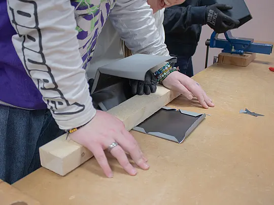 Zwei Schülerinnen arbeiten gemeinsam an einem Aluminium-Vogelhaus. Eine der Schülerinnen nimmt ein Holzstück, um das Aluminiumblech zu stabilisieren und zu formen, damit es die richtige Form für das Häuschen erhält. Beide konzentrieren sich auf die präzise Bearbeitung des Blechs. Auf dem Tisch sind Teile sichtbar, die vom Blech abgeschnitten wurden.