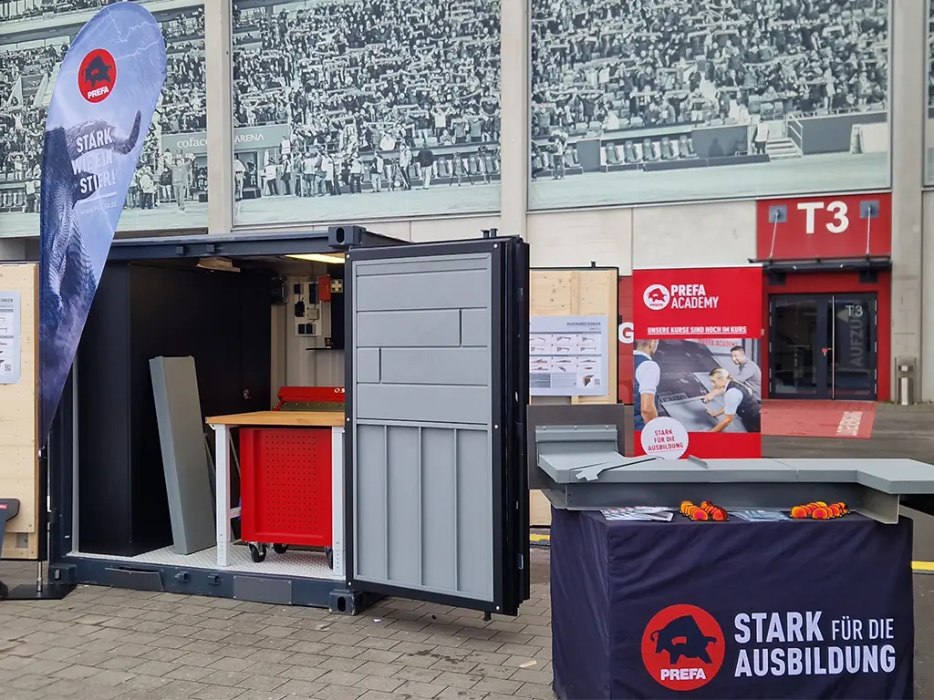 Mobiler PREFA Academy Cube auf einer Veranstaltung: geöffneter Container mit Werkbank, Werkzeugschrank und Fassadenmustern. Vor dem Container steht ein Präsentationstisch mit PREFA Branding und Bauteilen für Dach- und Fassadensysteme, im Hintergrund ein PREFA Academy Messestand.