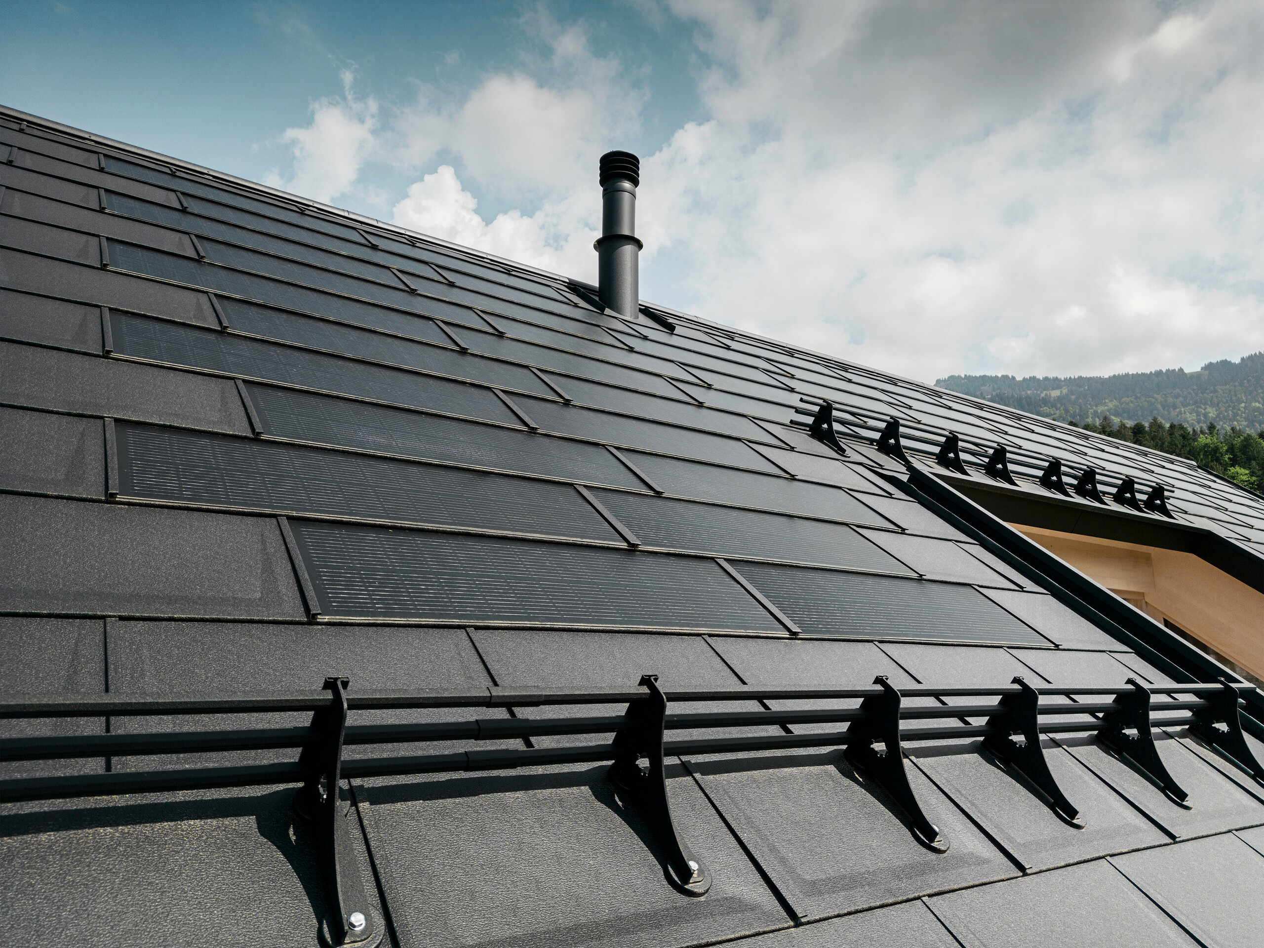 Nahaufnahme der effizienten PREFA Solardachplatte, welche sich nahtlos integriert in das schwarze Dach aus robusten PREFA Dachplatten R.16. Die Kombination aus nachhaltiger Energiegewinnung und langlebigem Aluminiumdach bietet eine ökologische und ästhetische Dachlösung, die sich harmonisch in die natürliche Landschaft einfügt.