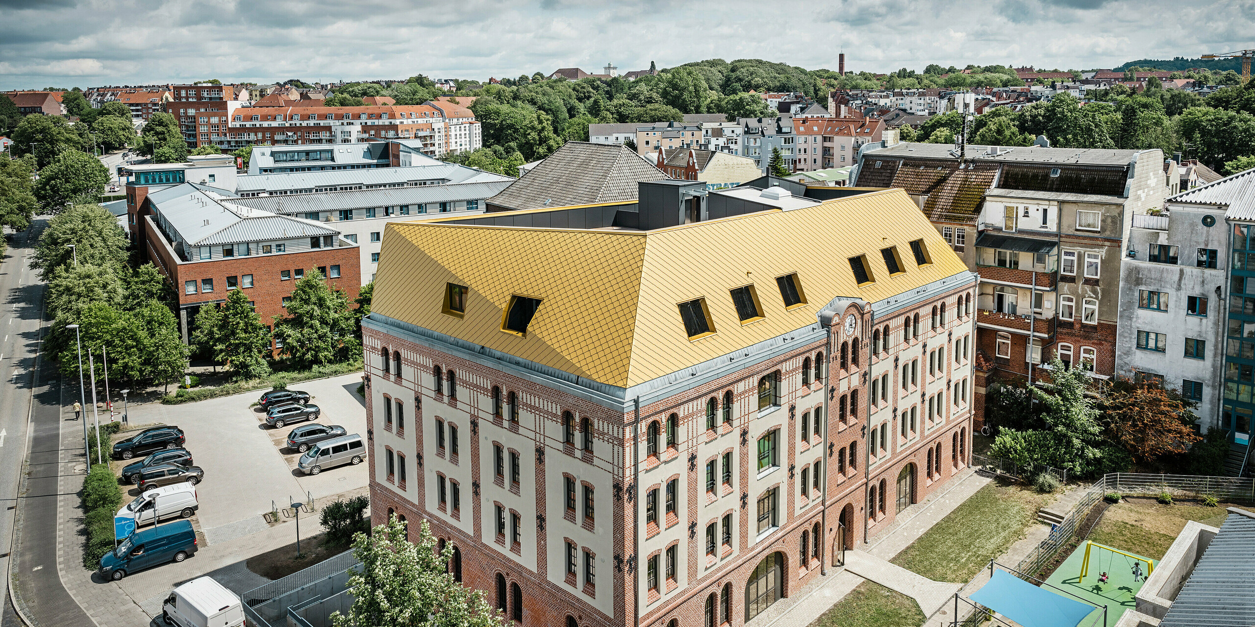 PREFA Dachrauten und Wandrauten 29 × 29 in Mayagold auf der Spitze des Studentenwohnheims in Kiel. Das Gebäude ist aus der Vogelperspektive mit einem Stadtpanorama im Hintergrund zu sehen. 