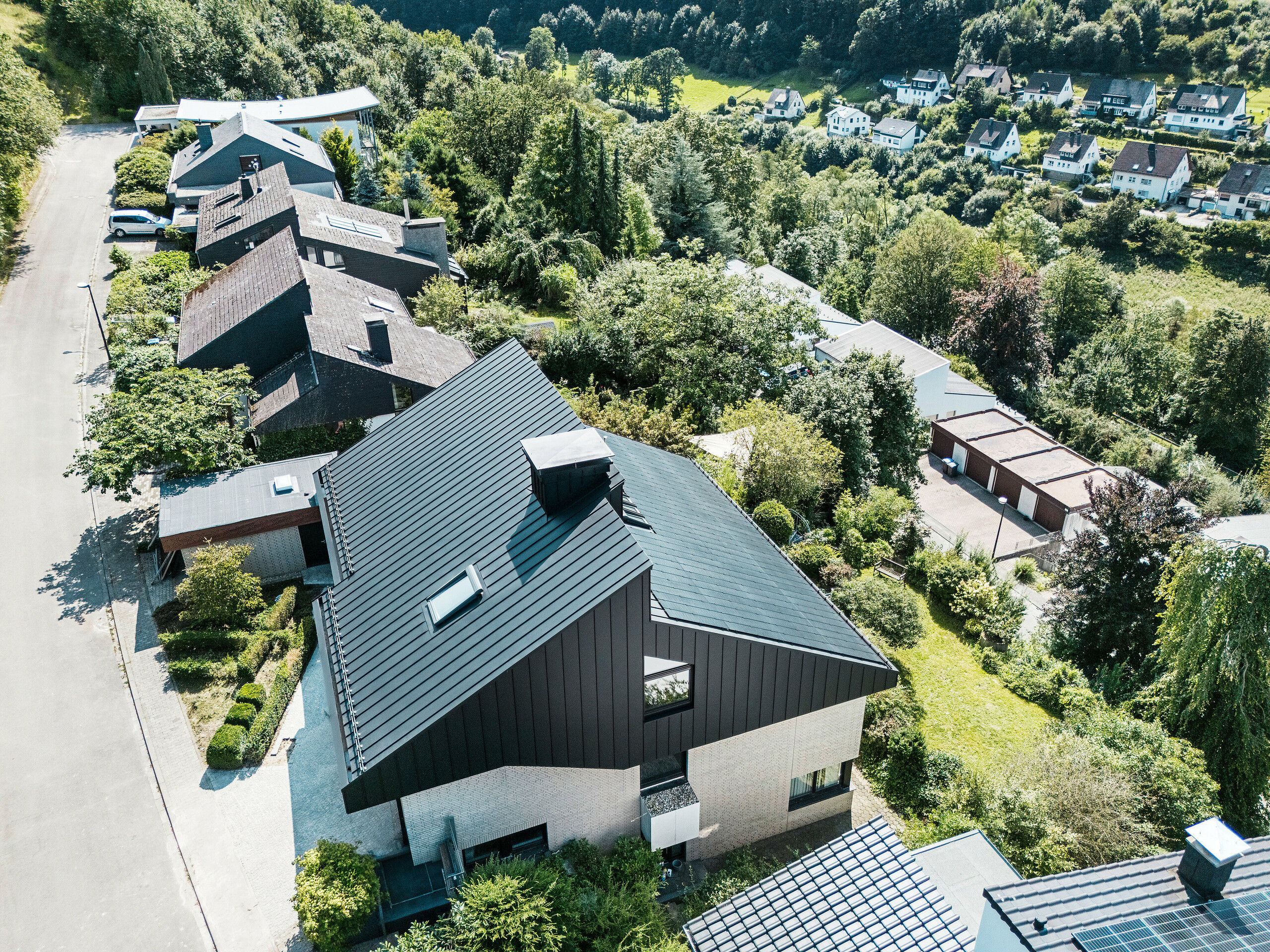 Einfamilienhaus in Meschede, Deutschland, mit versetztem Pultdach und innovativen PREFA Aluminiumprodukten. Die Dachfläche kombiniert eine formschöne Doppelstehfalzdeckung aus PREFALZ P.10 schwarz und rahmenlose Glas/Glas PV-Module mit TOPCon Technologie zur umweltfreundlichen Stromerzeugung aus erneuerbarer Energie. Die Aluminiumfassade an der Giebelseite und die nachhaltige Photovoltaik-Aufdachanlage unterstreichen die moderne Architektur, Funktionalität und Nachhaltigkeit.