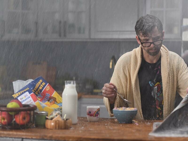 Mann im Morgenmantel sitzt am Frühstückstisch mit einer Zeitung. Es regnet im Haus und alles ist nass. Ein Einblick in den neuen PREFA Werbespot.
