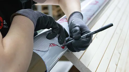 Ein Meisterschüler mit Arbeitshandschuhen markiert mit einem Stift eine Linie auf einem gebogenen PREFA Aluminiumblech. Das Werkstück liegt auf einem Dachmodell in einer Werkstattumgebung – Teil der praktischen Übung im Klempnerhandwerk.