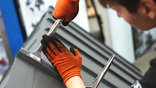 Ein Teilnehmer bearbeitet präzise eine Falzverbindung an einem Aluminiumdachmodell. Er trägt orange-schwarze Arbeitshandschuhe und nutzt spezialisierte Klempnerwerkzeuge, um das Material zu formen. Die Szene zeigt konzentrierte Handwerksarbeit im Rahmen des Trainings für die Deutsche Meisterschaft im Klempnerhandwerk.