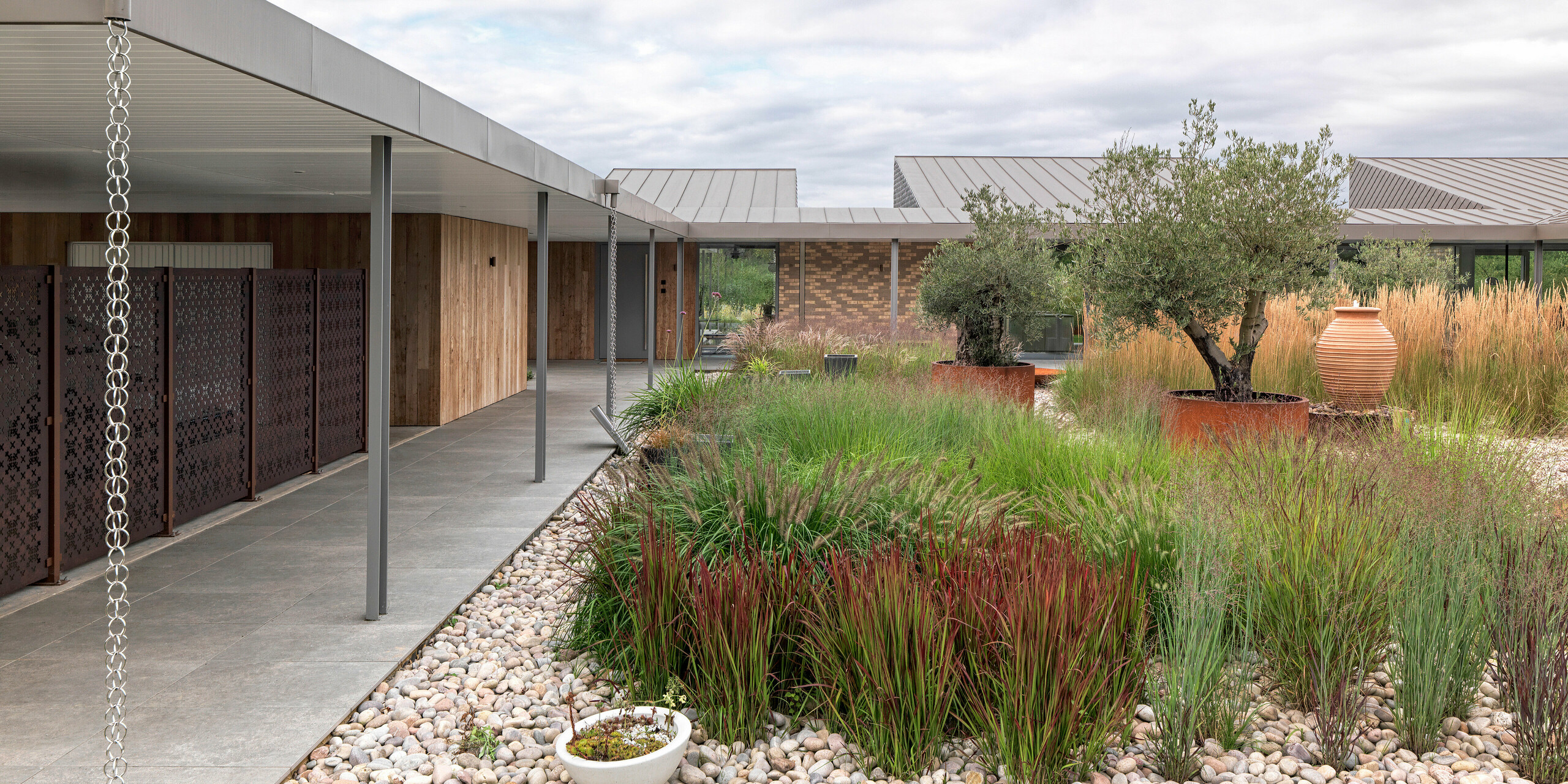 Detailansicht des modernen Wohnhauses in West Sussex mit Flachdach aus PREFALZ Aluminium in P.10 Bronze. Zu sehen ist die überdachte, klar strukturierte Terrassenanlage mit filigranen Säulen, stilvoll integrierten Regenablaufketten und einer abwechslungsreichen Gartengestaltung mit Ziergräsern, Olivenbäumen und Natursteinen. Die langlebigen PREFA Dachprodukte aus Aluminium unterstreichen die moderne Linienführung und fügen sich harmonisch in das nachhaltige Architekturkonzept ein.